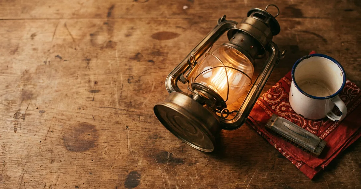 A brass porch lantern glowing on a worn wooden table beside an enamel coffee cup and a harmonica, illustrating a guide to how to write a country song