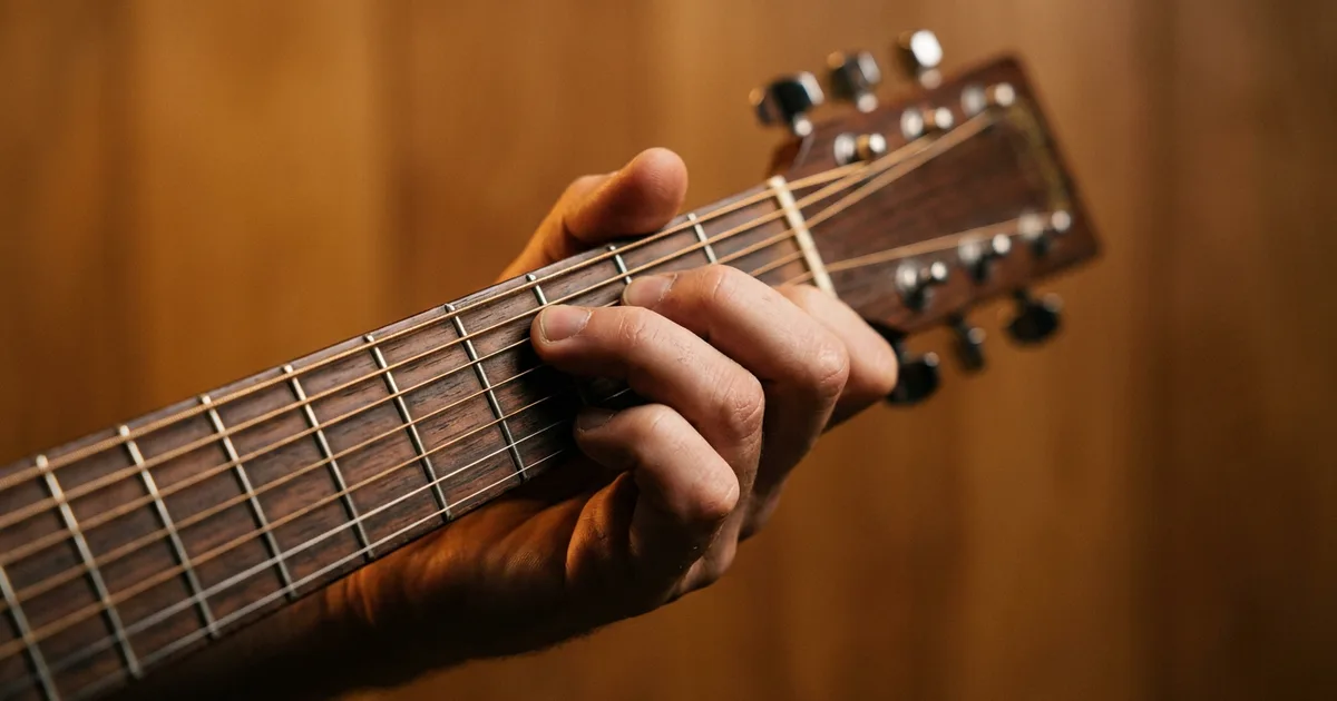 Writing a chord progression on guitar: a songwriter's hand forming a chord shape on the neck of an acoustic guitar in warm amber light.