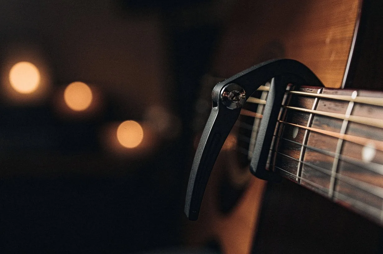 Black metal capo clamped across the second fret of an acoustic guitar neck, the strings pressed firmly behind the capo bar, warm amber side-light catching the chrome screw of the capo and the bronze of the strings, deep umber bokeh behind the neck