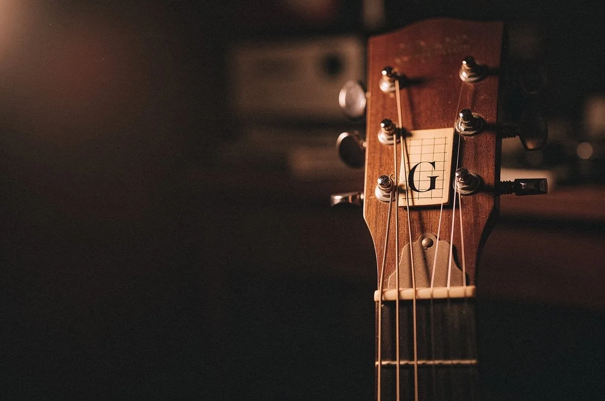 Acoustic guitar headstock and the first frets viewed from a low angle, the tuning pegs catching warm copper side-light, the steel strings glinting against a dark umber background, intimate songwriter workspace atmosphere with deep bokeh fading to near-black behind