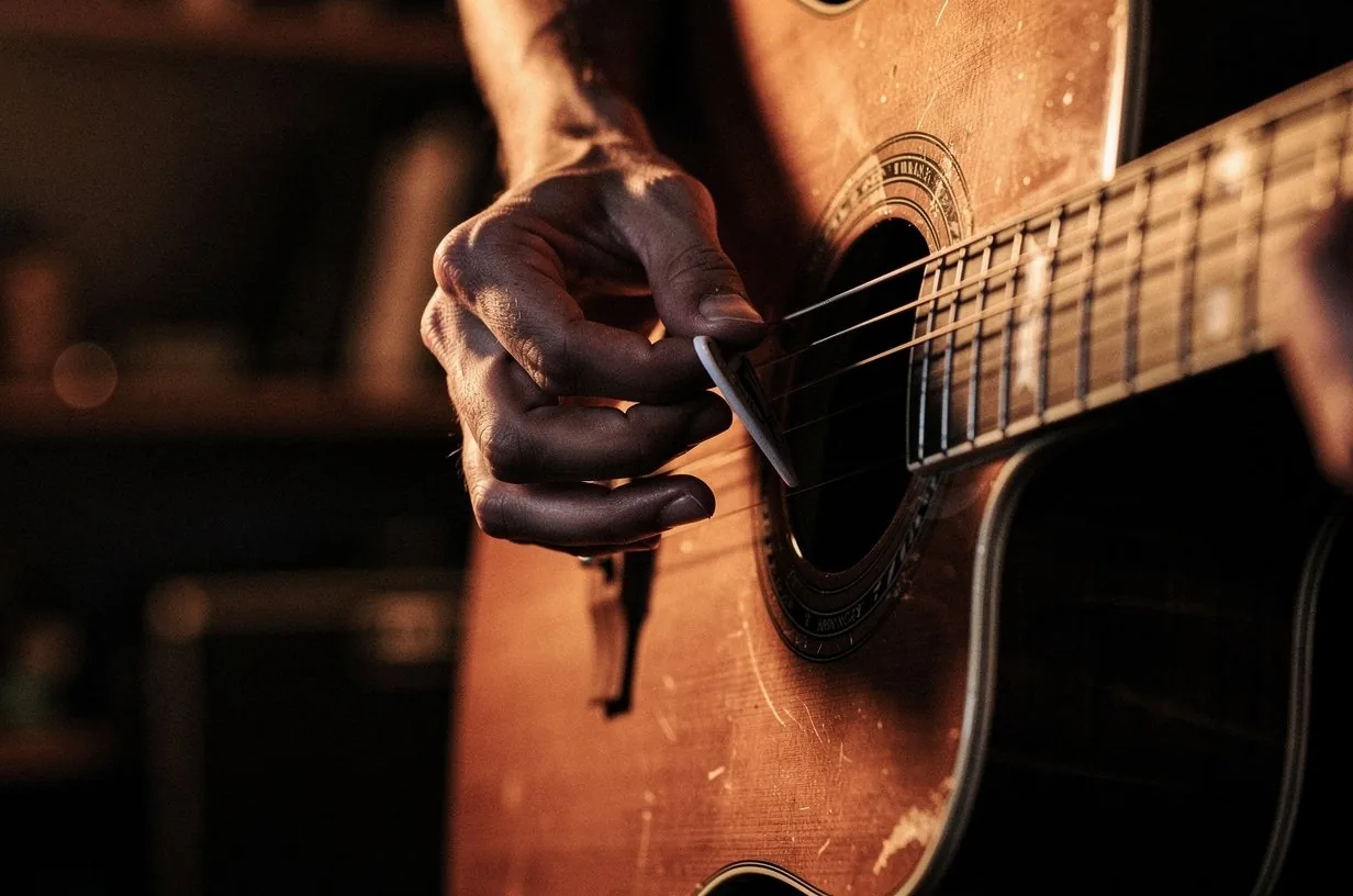 Right hand strumming an acoustic guitar pick across the strings near the soundhole, the strings catching warm amber light, the wood grain of the rosette visible in sharp focus, motion blur on the pick as it sweeps downward, intimate songwriter atmosphere
