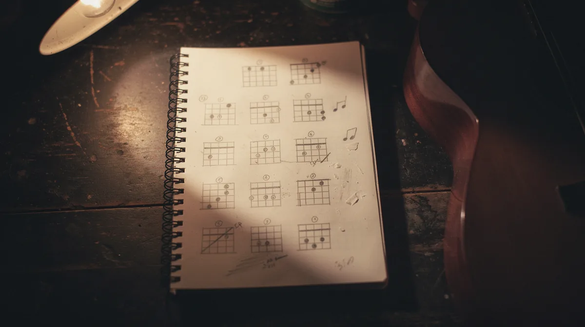 Overhead view of an open spiral-bound notebook on a dark wooden desk filled with hand-drawn guitar chord diagrams in pencil arranged in a grid, the rosette and edge of an acoustic guitar entering the right side of the frame, a small desk lamp at the upper-left throwing a warm pool of light onto the page, the rest of the desk fading into deep amber-brown shadow, intimate songwriter workspace