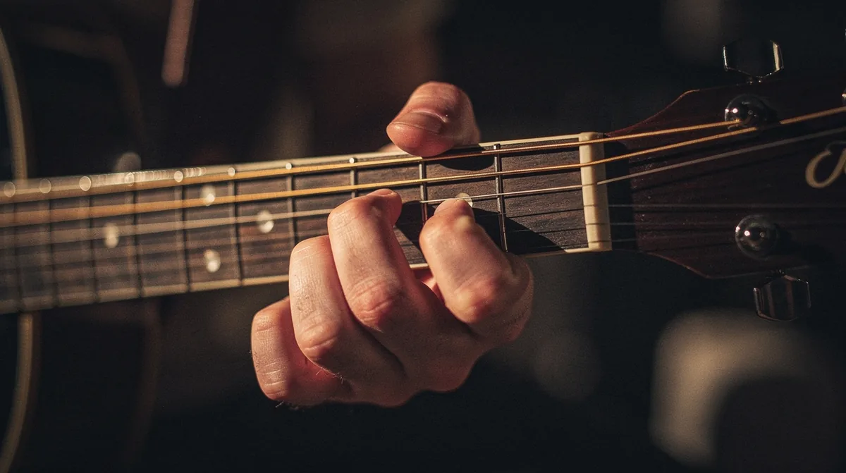 Close-up of a guitarist's left hand high on the neck of an acoustic guitar around the twelfth fret with a single index finger pressing a single string for a lead-line note, the rest of the fretboard receding into deep bokeh, warm amber rim-light catching the steel strings, dark umber-brown background, shallow depth of field