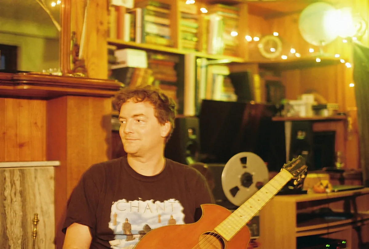 Songwriter Paul Spring in a black t-shirt holding his 12-string guitar, with a reel-to-reel tape machine and bookshelves behind him