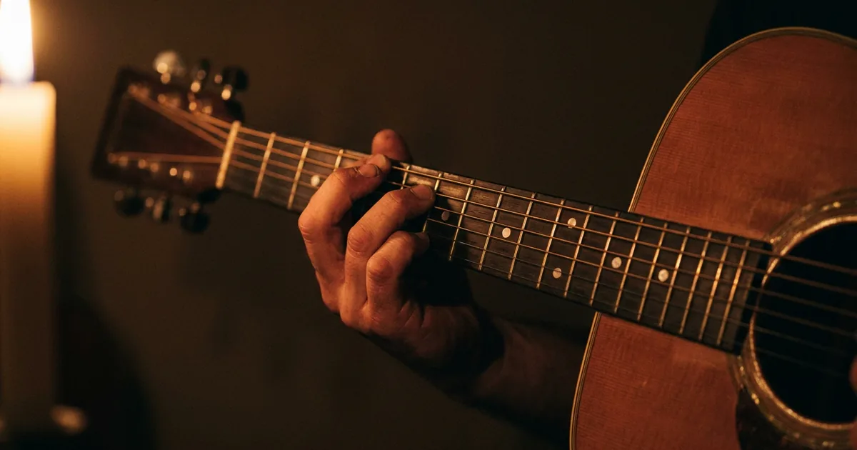 A hand forming an augmented chord shape on an acoustic guitar fretboard in warm amber light