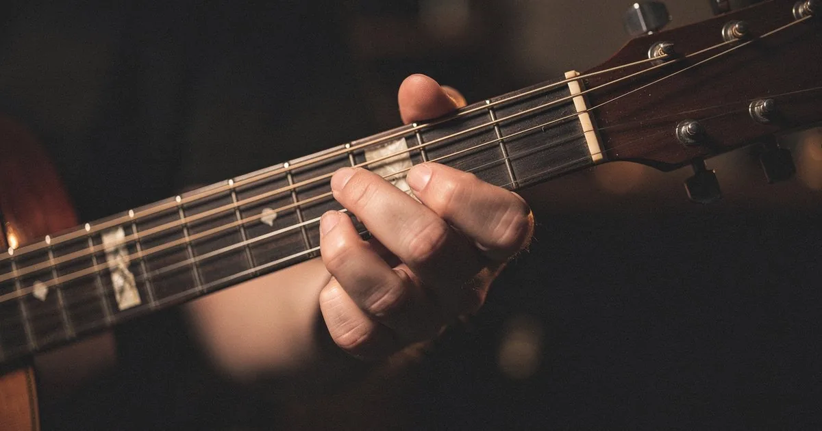 A hand pressing a chord shape on an acoustic guitar fretboard in warm amber light, the kind of voicing a songwriter reaches for when writing in a musical mode.