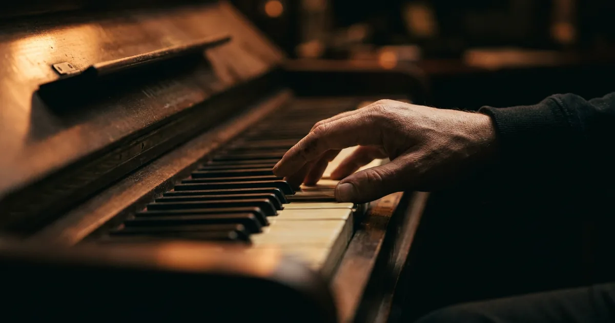 A hand poised over a narrow span of piano keys, the rest of the keyboard in shadow, illustrating a songwriting exercise that limits the melody to a few notes