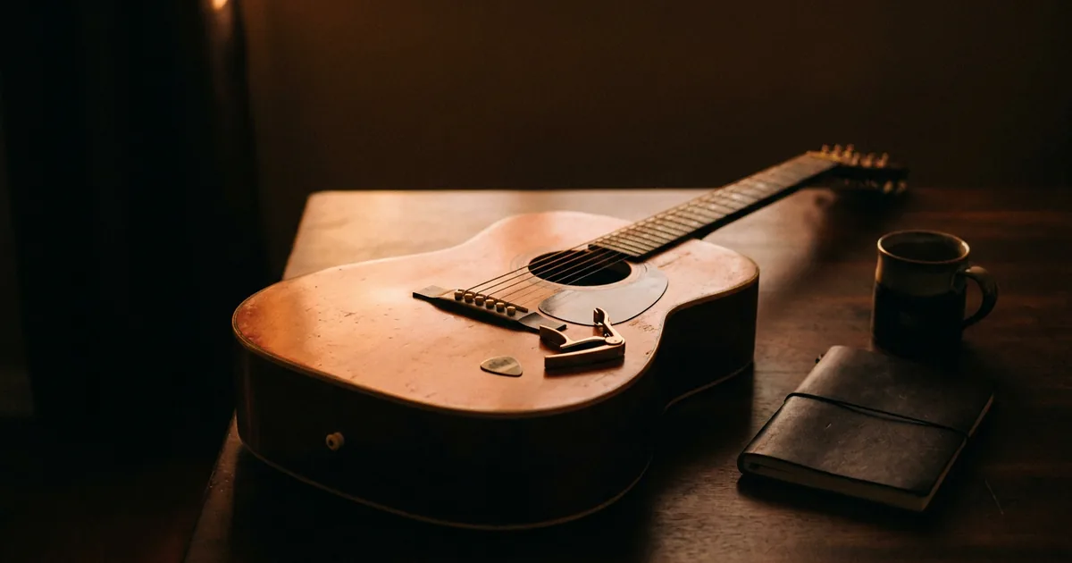 An acoustic guitar resting on a dark table with a capo and pick on the soundboard and a closed notebook beside it, the setup a songwriter uses to test whether a chorus lifts out of a verse